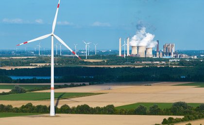 A wind turbine in a dry field with a power plant emitting steam in the background