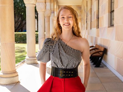 A young woman with long blonde hair standing under a sandstone walkway surrounded by columns