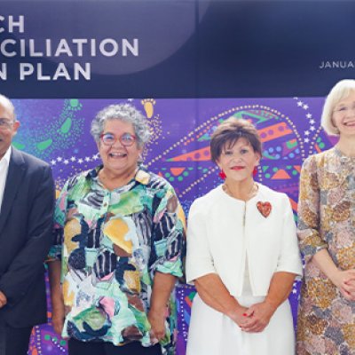 A man and three women stand on a stage in front of a UQ banner that reads Stretch Reconciliation Action Plan. 