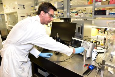 A man in a white labcoat, goggles and gloves works on a machine in a laboratory.