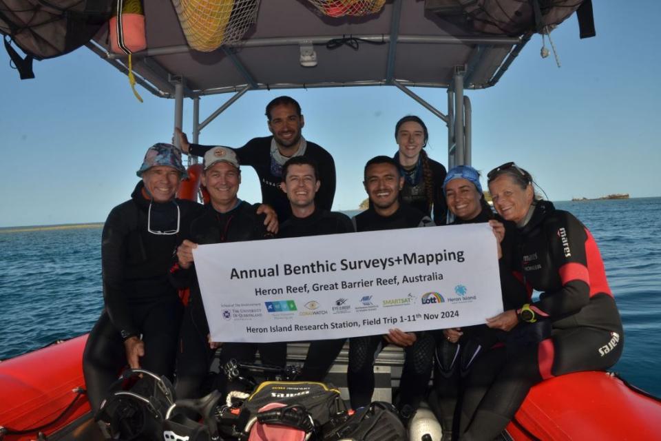 Eight divers sitting on a boat holding a sign stating the annual benthic surveys and mapping