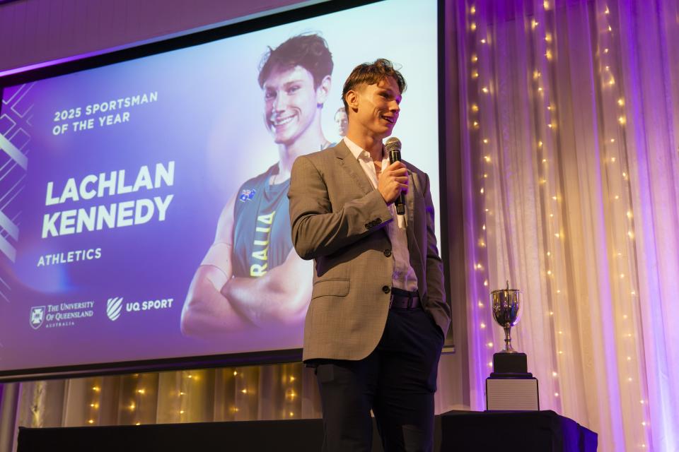 Lachie Kennedy holding a silver trophy while wearing a grey sports coat standing in front of a purple backdrop.
