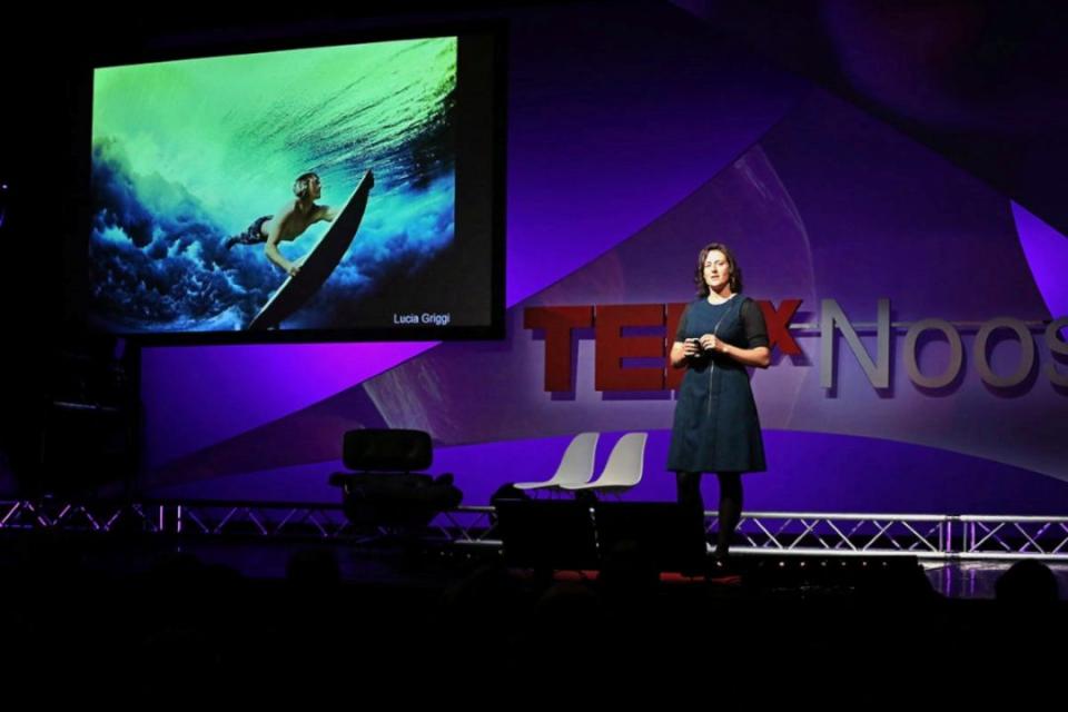A woman with brown hair wearing a dress presenting on stage for TED talks