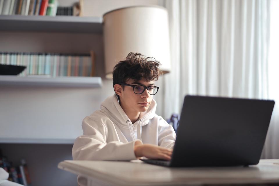 Yong boy with gallsess and white jumper uses a laptop on a table