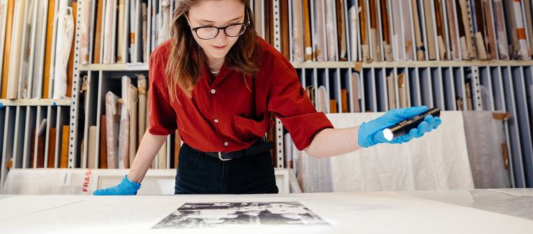 A woman in a red shirt, wearing blue latex gloves, uses a torch to look closely at an archival photo on a desk. 
