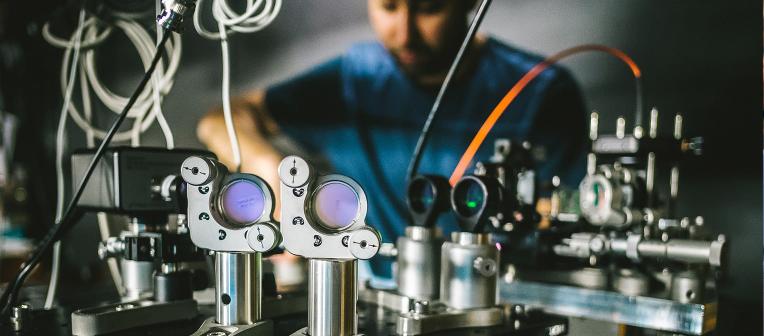 A complicated-looking machine is in focus in the foreground, with a person in the background making adjustments to it.