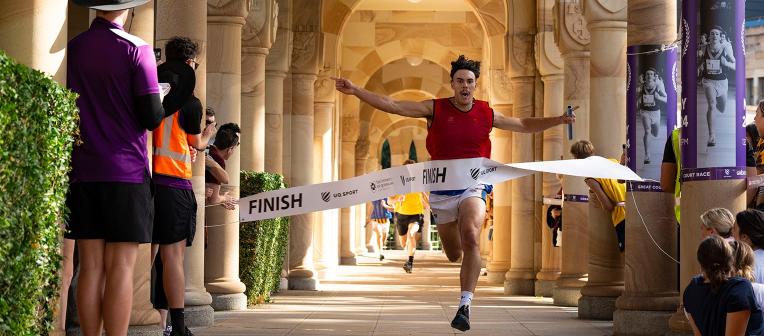 A man, holding a baton and running in the UQ Cloisters as part of the Great Court Race, breaks through the finish line tape.