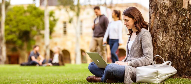 A woman sits under a tree in the UQ Great Court, reading the news on her laptop