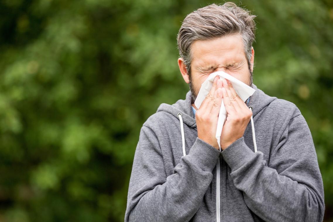 A man sneezes into a tissue as he stands outside in front of trees.