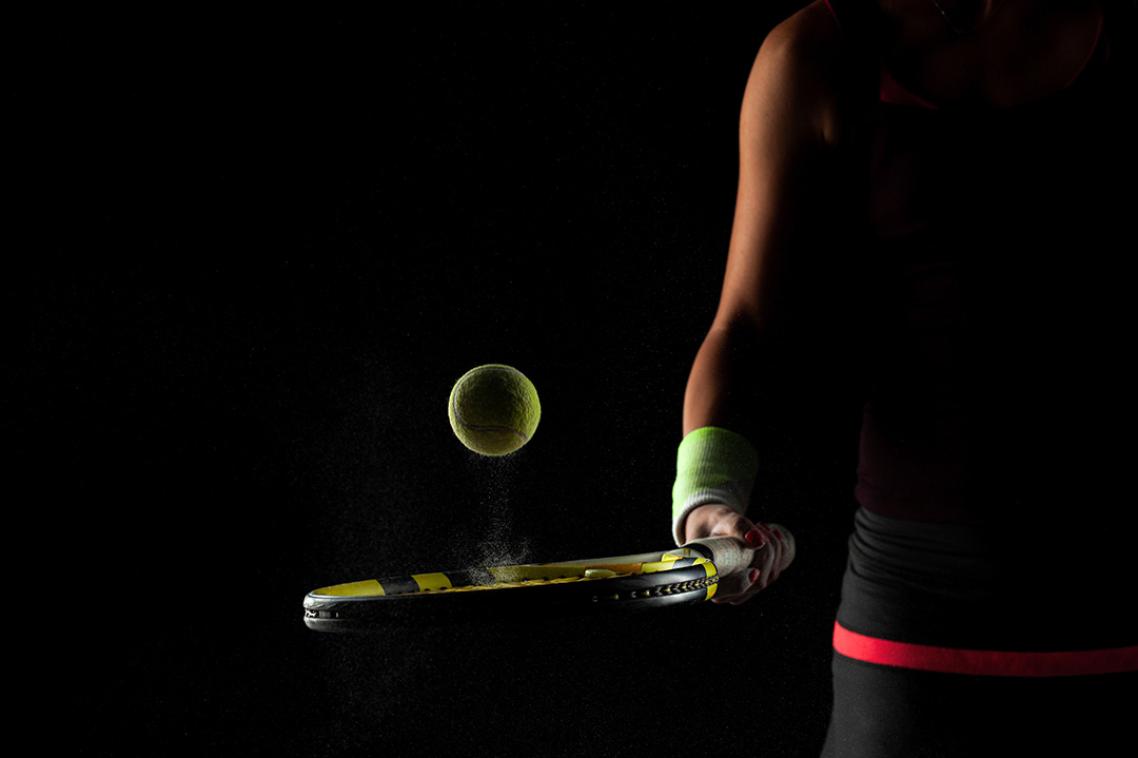 A sports person bounces a tennis ball off a racquet against a black background.