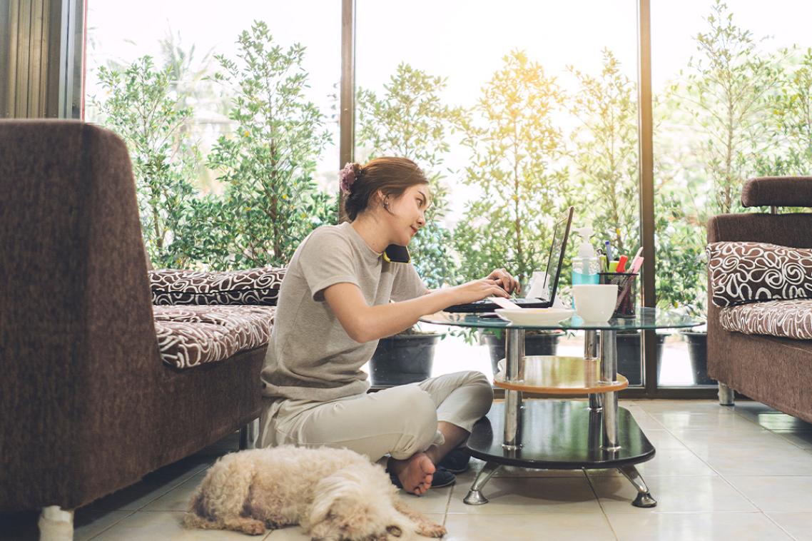 A woman is working from a loungeroom, balancing a phone and laptop on a coffee table.