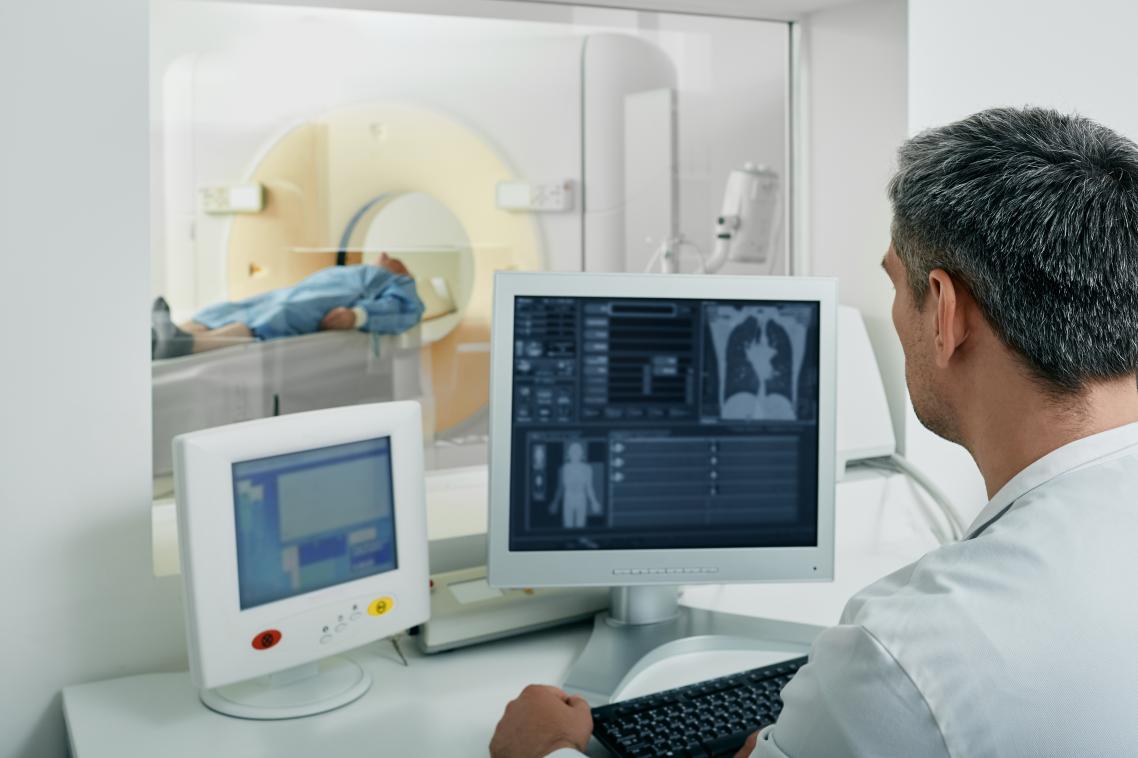 Man in white coat looking at radiology images with patient in CT scanner in background. Adobe