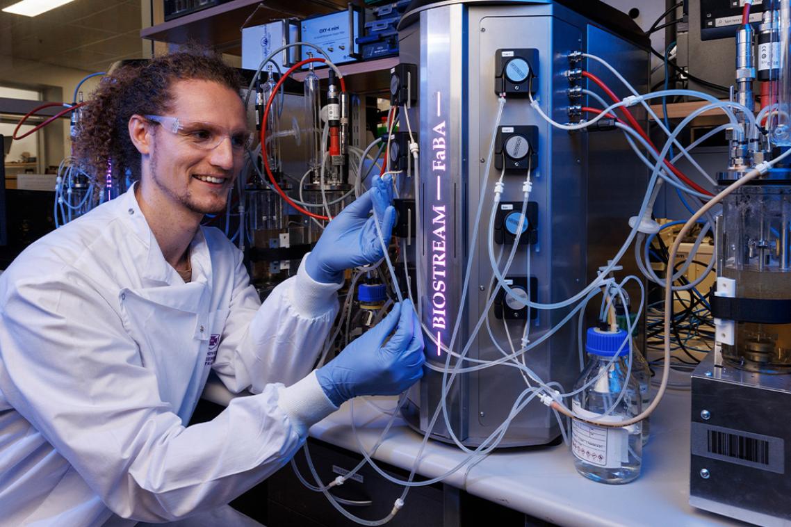 A male scientist in a white coat, goggles and gloves sites next to a machine with lots of tubes and lights.