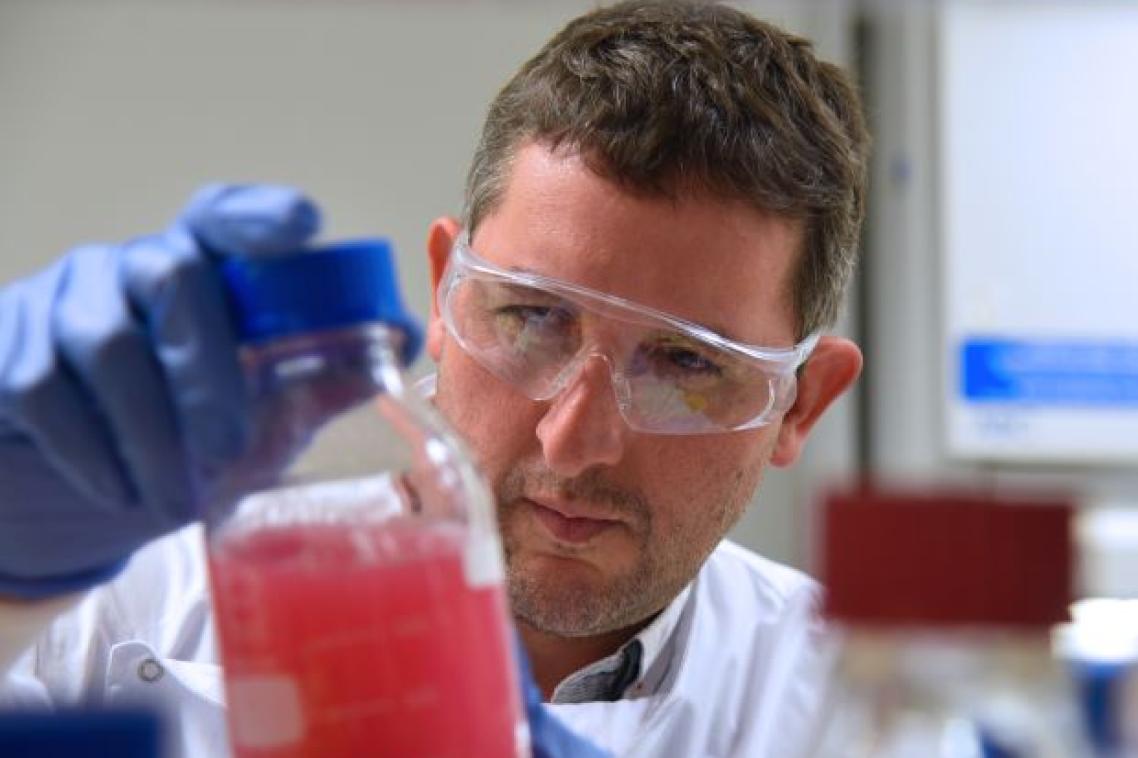 A male scientist in lab coat and goggles inspects a glass full of red liquid. 