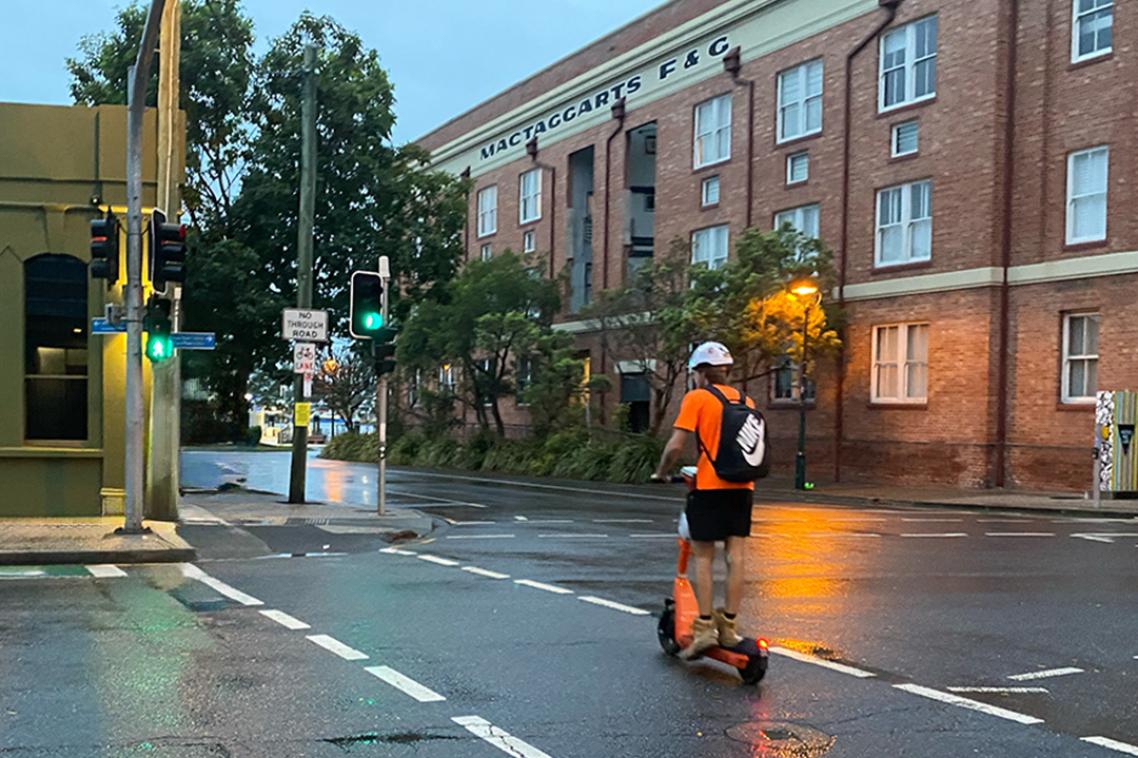 A person in high-vis and helmet rides an e-scooter across a road on a rainy day.