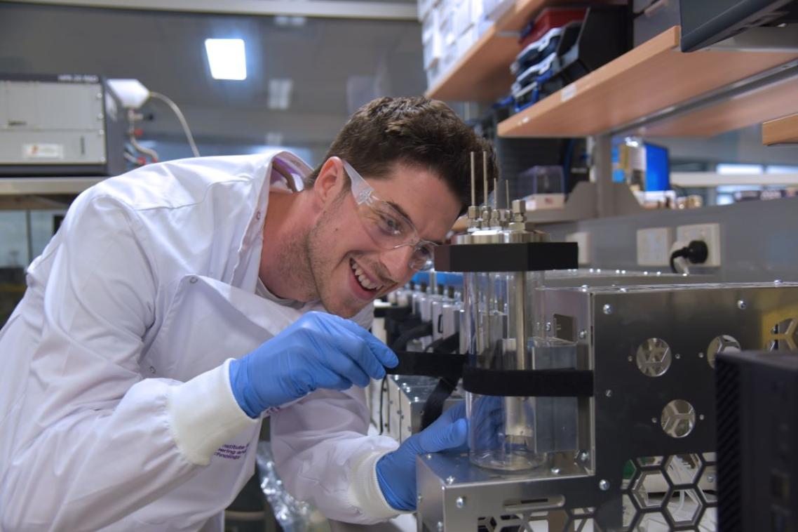 a man in a white lab coat and glasses adjusts a glass cylinder surrounded by stainless steel equipment