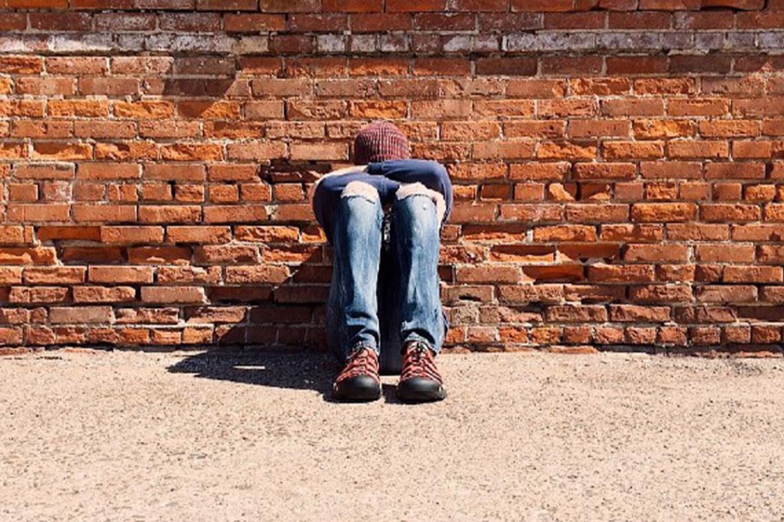 A young person sits against a brick wall with their head down.