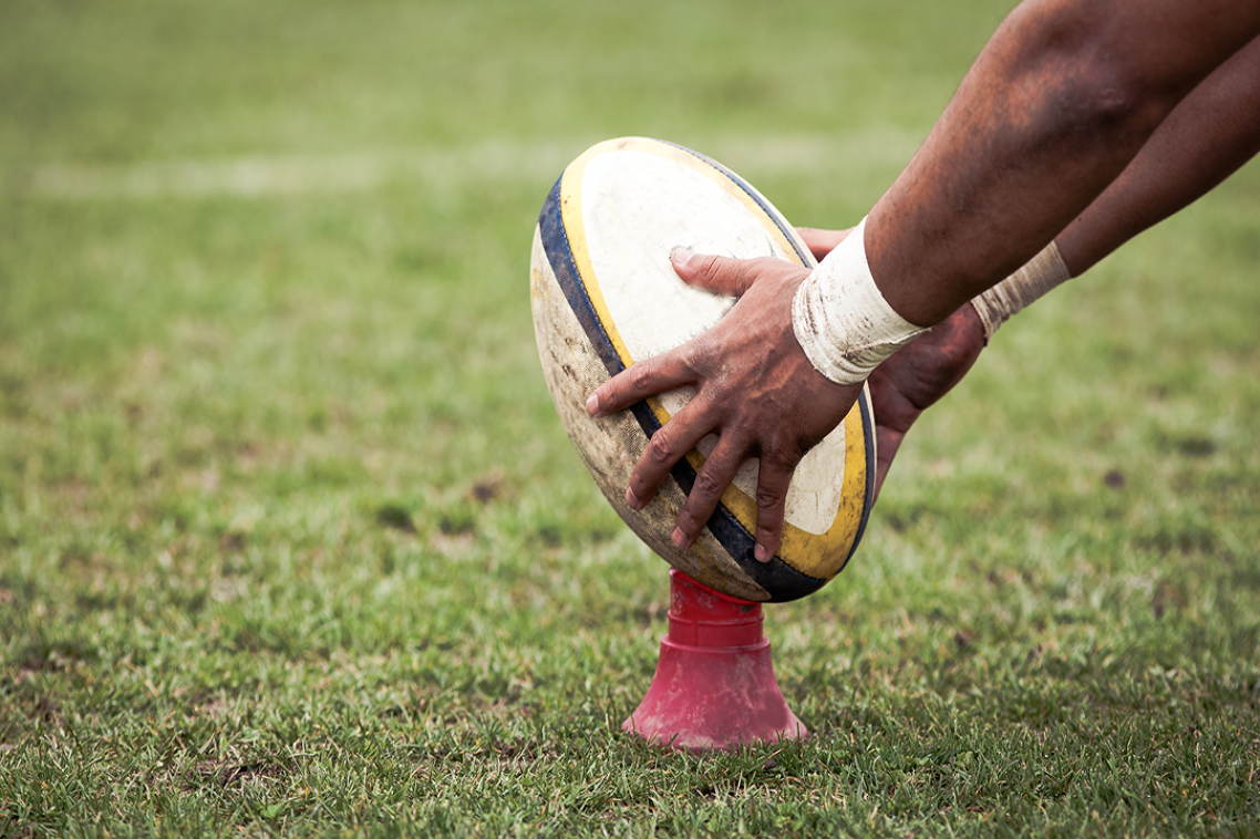 Hands holding a white rugby ball on a red kicking tee.
