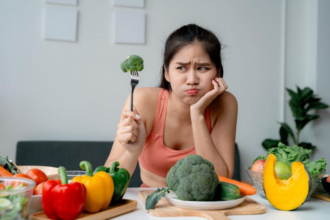 An unimpressed woman holding up a piece of broccoli on a fork.