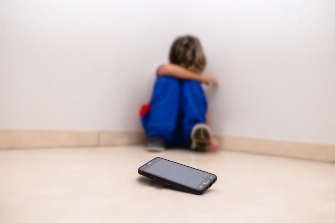 An upset child huddled in a corner who's thrown his phone on the floor. 