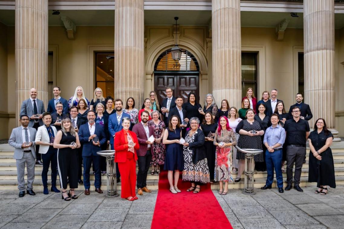 Large group of men and women standing before a grand building with sandstone columns and a red carpet.