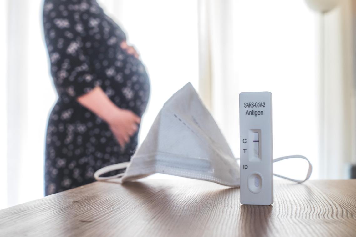 A positive COVID-19 test and mask in the foreground, with a pregnant woman standing in the background.