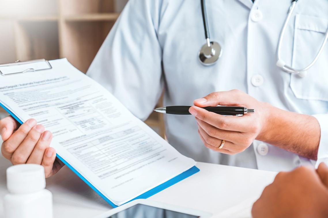 A person with a stethoscope and holding a pen and clipboard with medication on a desk is doing a consultation with a patient.
