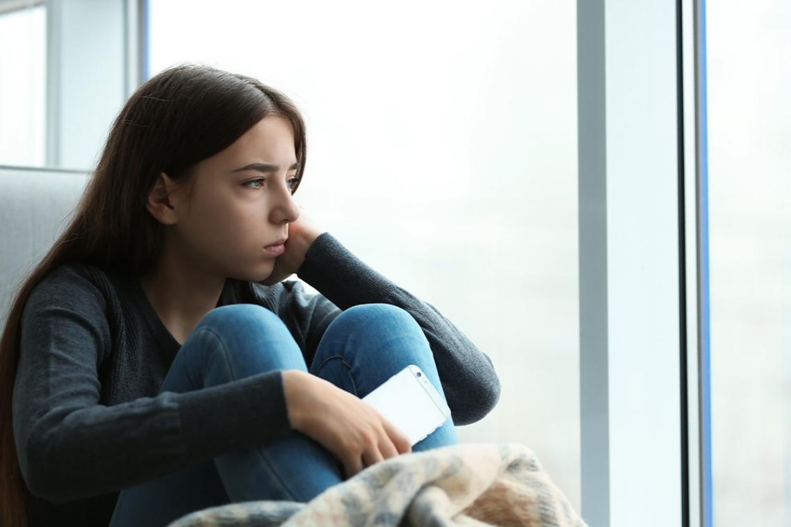 A young girl holding a mobile phone and sitting by a window looks sad.