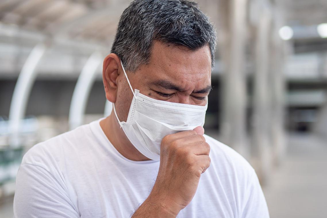 A man wearing a white shirt and a white medical mask coughing into his hand.