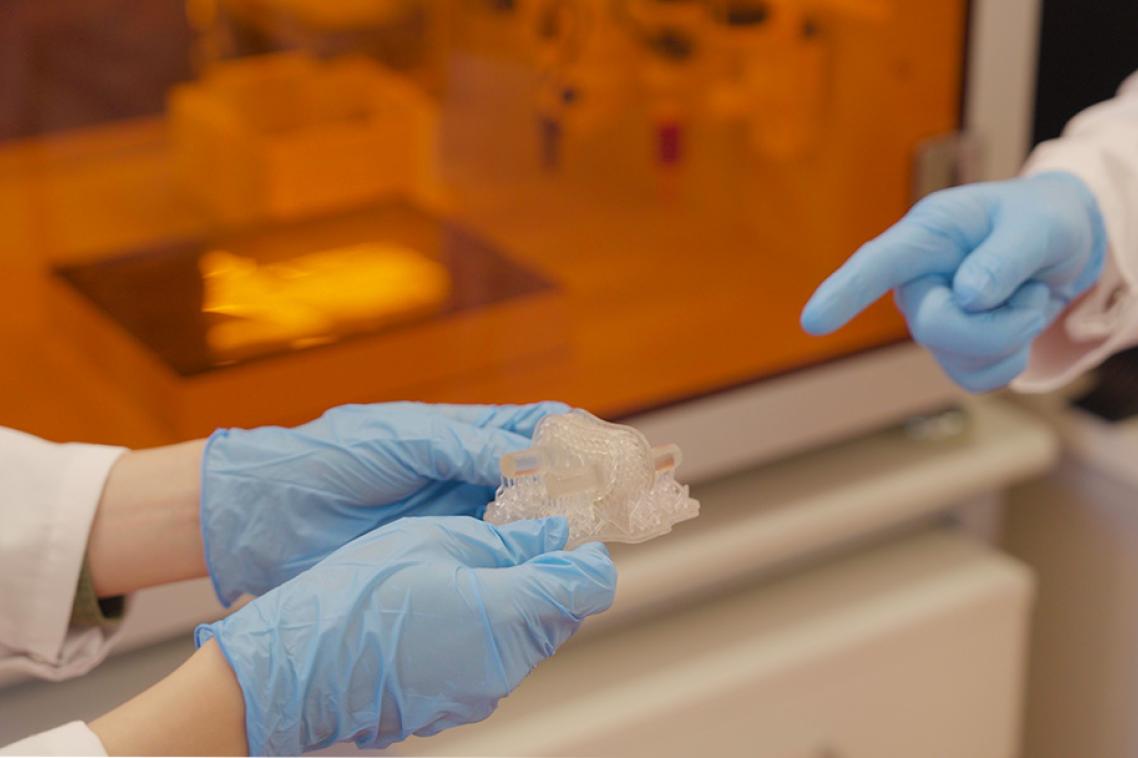 Two gloved hands holding a small 3D printed device in front of a incubator