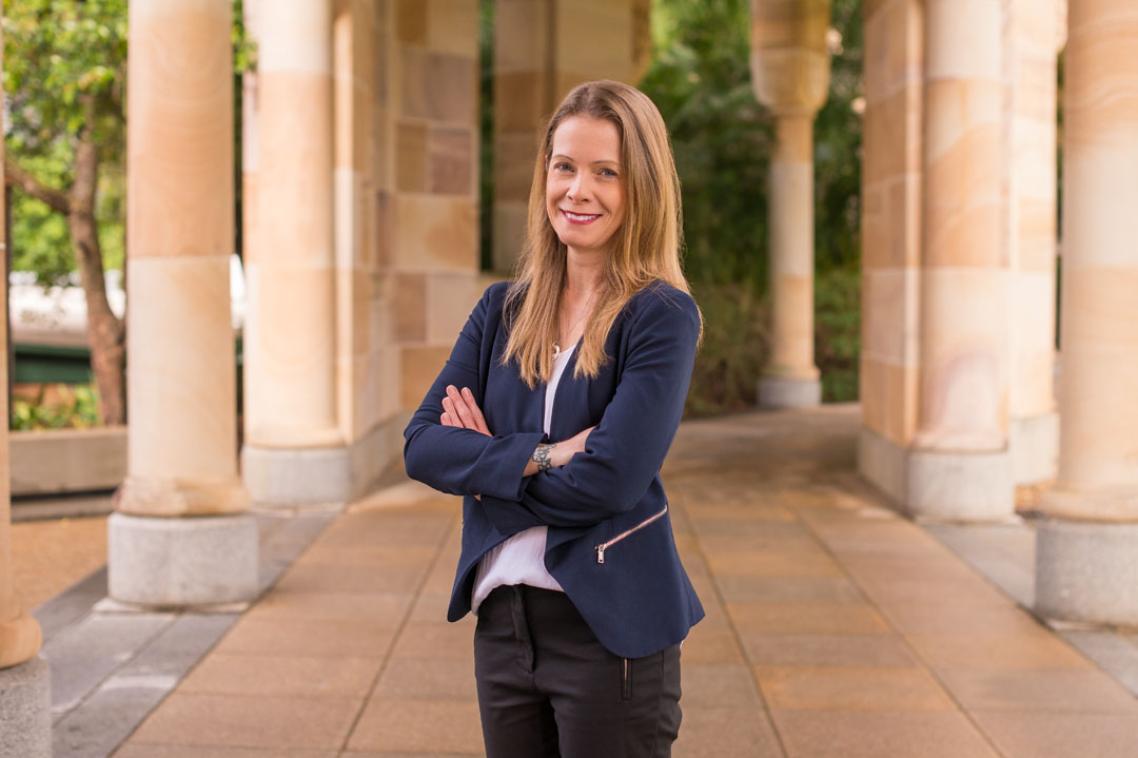 A woman in a blue blazer standing with her arms crossed in a sandstone corridor