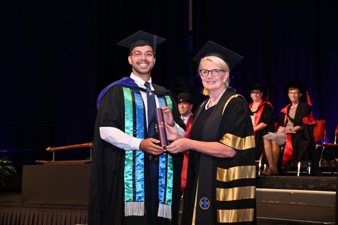 A man and woman in graduation gowns, the man holding a purple scroll