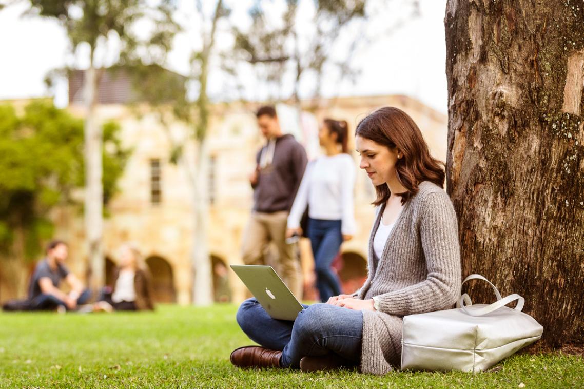 A woman sits under a tree in the UQ Great Court, reading the news on her laptop