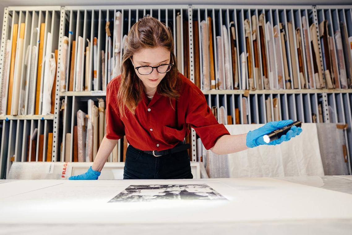 A woman in a red shirt, wearing blue latex gloves, uses a torch to look closely at an archival photo on a desk. 