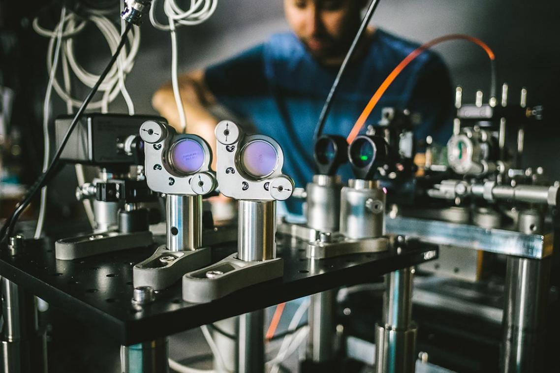 A complicated-looking machine is in focus in the foreground, with a person in the background making adjustments to it. 