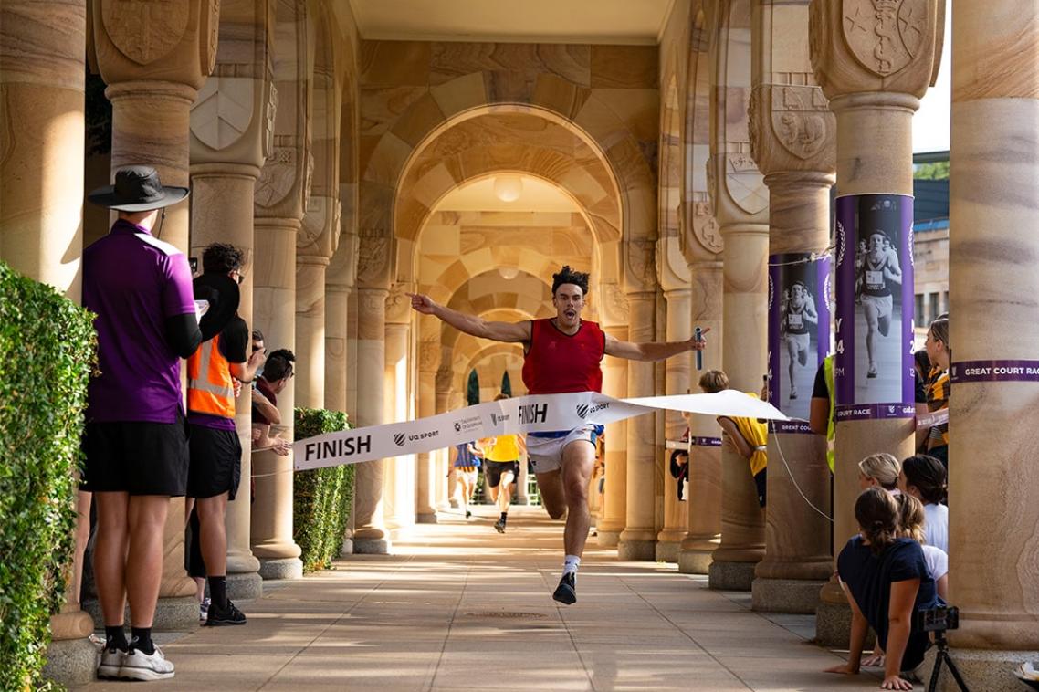 A man, holding a baton and running in the UQ Cloisters as part of the Great Court Race, breaks through the finish line tape. 