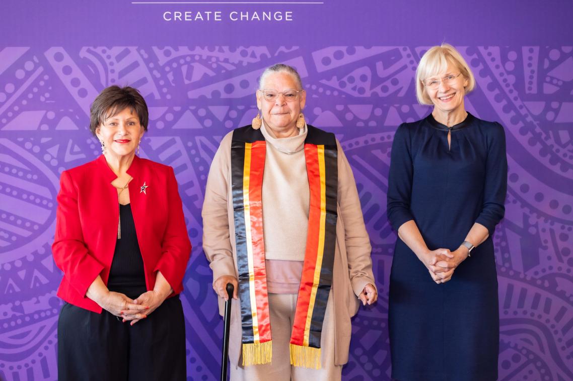 Three women standing in front of a purple backdrop smiling to camera.
