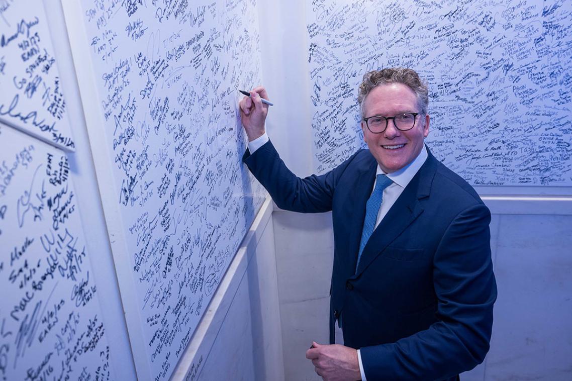 A man in a blue shirt writing on a wall and staring at the camera