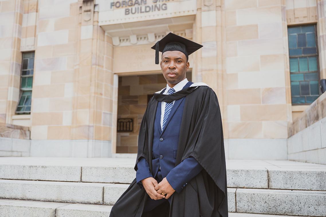 A young man stands in graduation cap and gown outside UQ's Forgan Smith building.