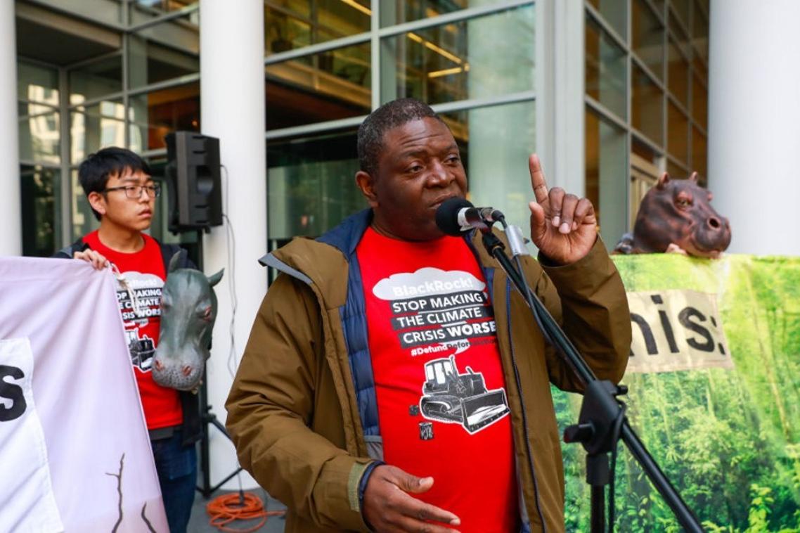 A man stands in front of a microphone at a demonstration with protest banners behind him.