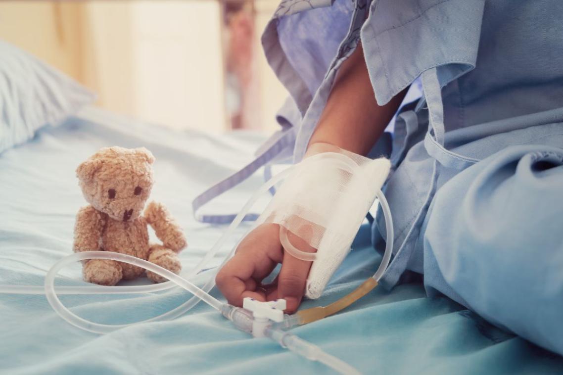 A child sitting on a hospital bed with an IV inserted and a teddy bear for comfort. 