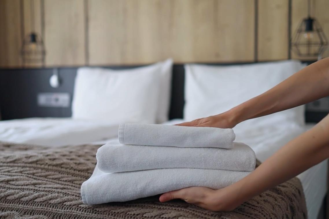 a pair of hands holds a stack of neatly folded towels above a hotel bed