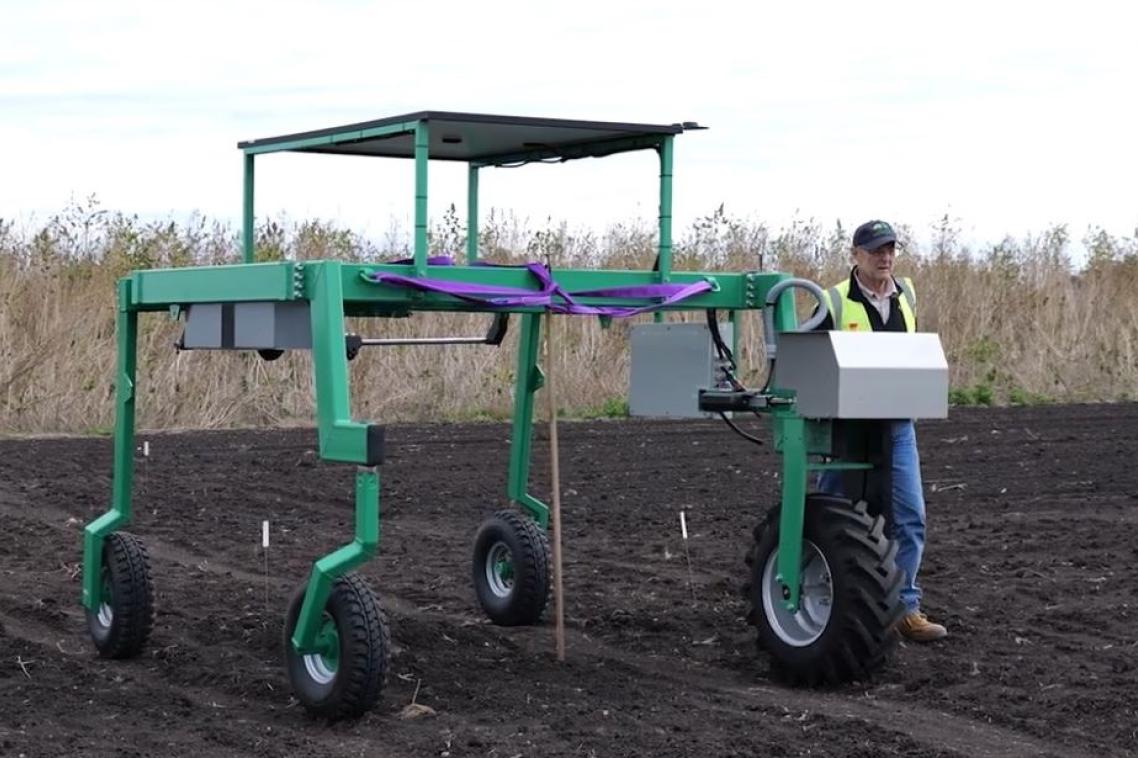 a man walks next to a 4-wheeled, tall green machine in a field