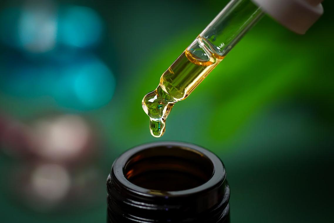 A droplet hovers on the end of a dropper over an open oil bottle, on a green and blue backdrop.