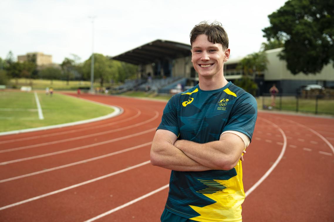 Lachie Kennedy in a green and gold Australia shirt standing on a red athletics track with his arms crossed, smiling at the camera.