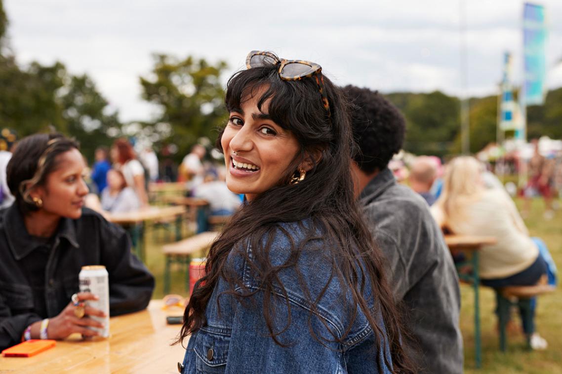 A young woman with long black hair and wearing a denim jacket turns smiling to the camera. She is sitting at an outdoor table with other people in the background. 