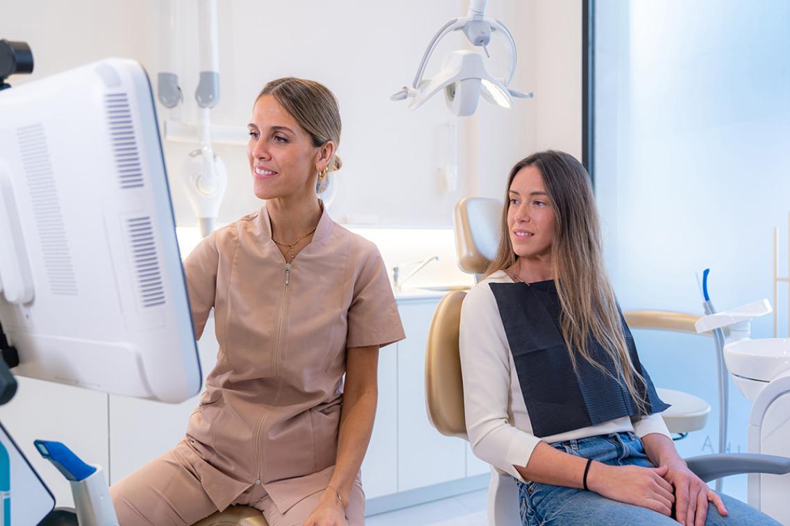 A dentist in pink scrubs pointing at a screen while a patient watches from behind.