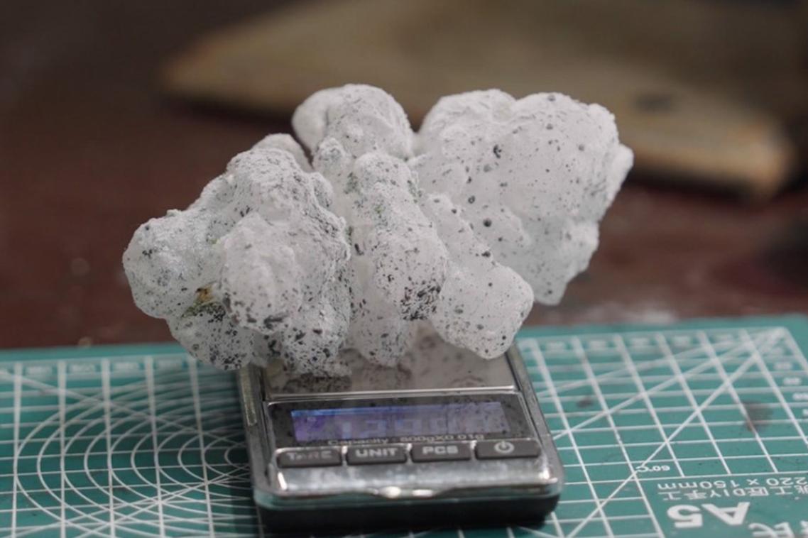A white spiky hailstone, flecked with black paint, sits on a set of digital scales.
