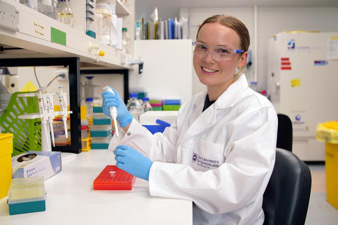 Dr Summa Bibby sitting in a lab holding a pipette while wearing a white lab coat and safety glasses.