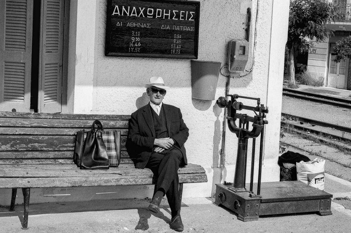 Black and white image of a man sitting on a park bench, in front of a sign written in Greek